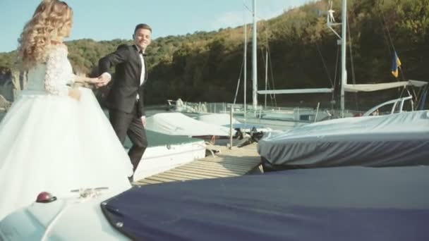 The groom is holding his bride by the hand. They are running on the pier. — Stock Video