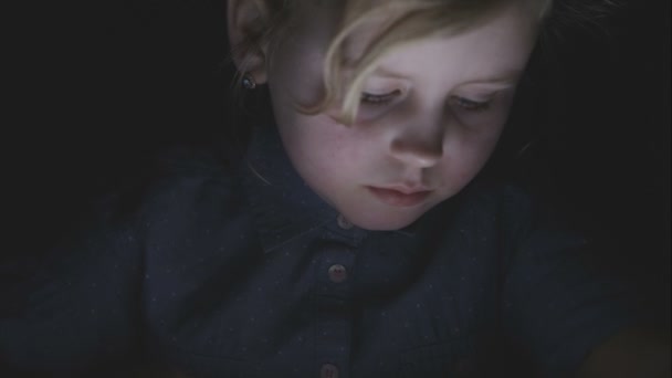 Gadis kecil melihat ke dalam tablet pc dalam gelap dengan pantulan cahaya di wajahnya — Stok Video
