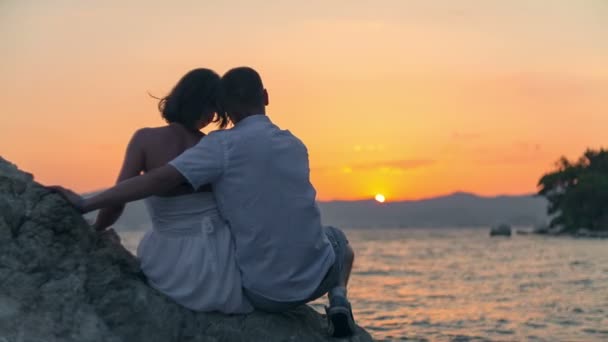 Casal jovem senta-se na rocha e espera pelo pôr do sol. Gravação de vídeo RAW . — Vídeo de Stock
