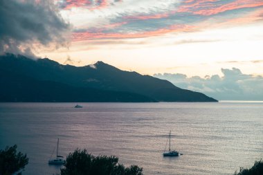Alacakaranlıkta sakin bir deniz manzarası. Dalgalı bir gökyüzü altında yelkenliler ve dağlar. Elba İtalya
