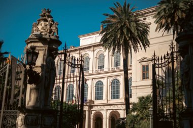 Mavi gökyüzünün altında palmiye ağaçları bulunan tarihi Avrupa binasında mimari ve demir kapılar yer alıyor. Roma İtalya