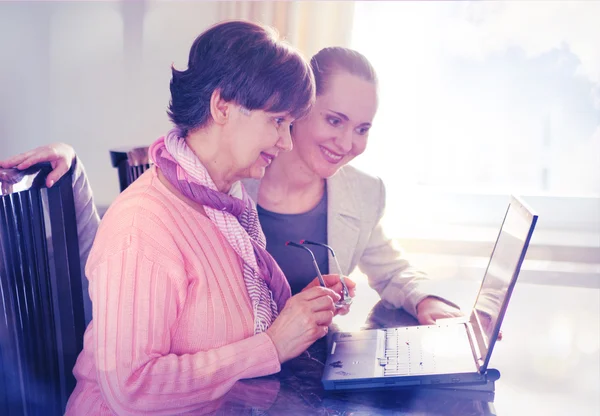 Młodsza kobieta pomaga osoby w podeszłym wieku przy użyciu komputera przenośnego dla wyszukiwarek internetowych. Młody i emerytury wiek pokoleń pracując razem. — Zdjęcie stockowe