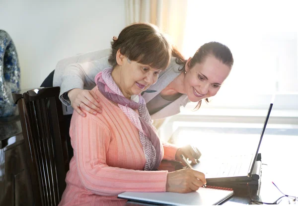 Yngre kvinna att hjälpa en äldre person med bärbar dator för sökning på internet. Unga och pension ålder generationer arbetar tillsammans. — Stockfoto