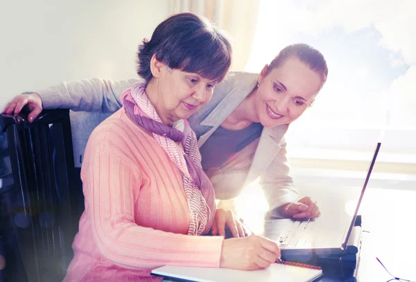 Mulher mais jovem ajudando uma pessoa idosa usando o computador portátil para pesquisa na internet. Jovens e gerações em idade de reforma a trabalhar em conjunto . — Fotografia de Stock