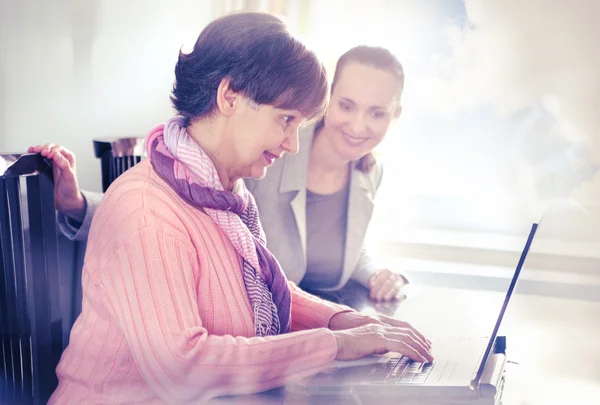 Yngre kvinna att hjälpa en äldre person med bärbar dator för sökning på internet. Unga och pension ålder generationer arbetar tillsammans. — Stockfoto