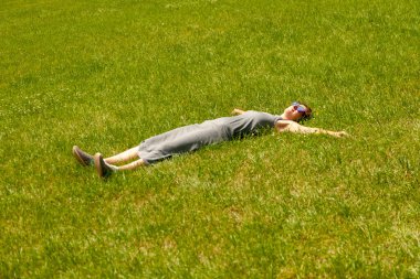 Yeşil çimen tarlasında gri elbiseli, spor ayakkabılı ve güneş gözlüklü genç bayan..