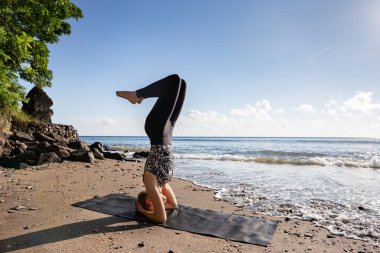 Siyahlı genç kadın Asya kumsalında yoga yapıyor Sirshasana (baş aşağı duruş) pozu. Sağlıklı yaşam tarzı, tropik bölgelerde mutlu tatiller, sabah egzersizleri..