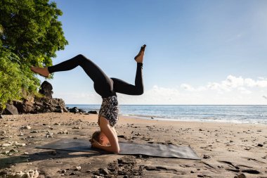 Siyahlı genç kadın Asya kumsalında yoga yapıyor Sirshasana (baş aşağı duruş) pozu. Sağlıklı yaşam tarzı, tropik bölgelerde mutlu tatiller, sabah egzersizleri..