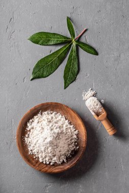 Cassava (manioc) flour in wooden bowl with original leaf on grey background top view. Alternative gluten free flour. Healthy food. clipart