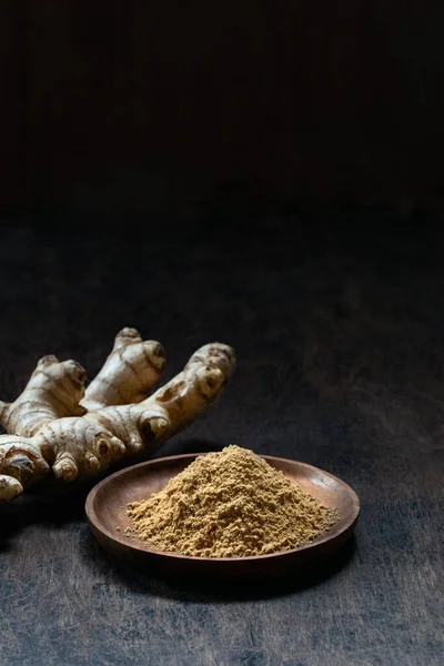 Fresh organic ginger root and powder in wooden bowl on dark old rustic background with copyspace vertical shot. Healthy food, diet, alternative medicine, Ayurvedic.