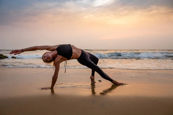 Middle age woman in black doing yoga on sand beach in India at sunset. Healthy lifestyle.