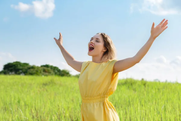 stock image Happy young woman in yellow dress is singing with closed eyes and raising her hands on green meadow. Enjoy summertime. Open your sound voise.