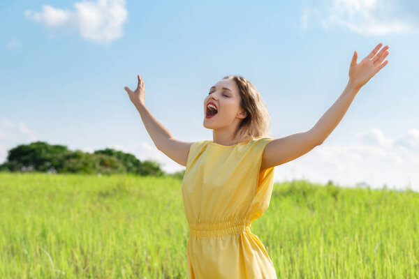 Happy young woman in yellow dress is singing with closed eyes and raising her hands on green meadow. Enjoy summertime. Open your sound voise.