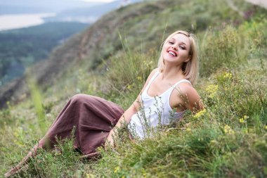 Güzel, gülümseyen sarışın genç bir kadın yeşil çimlerde oturuyor beyaz bluzlu ve kahverengi etekli tepe manzaralı.
