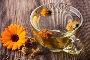 Calendula (Marigold) şeffaf cam bardakta bitkisel çay ve ahşap arka planda kurumuş çiçekler.