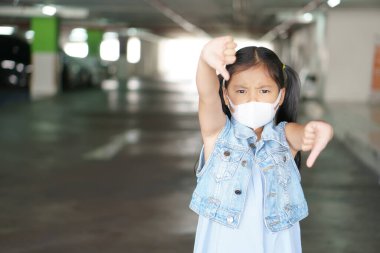 Asyalı çocuk başparmağı aşağıda veya çocuk N95 beyaz yüz maskesi takmaktan hoşlanmıyor nezle burnu hasta ve PM2.5 tozunu araba parkı dumanıyla veya covid-19 koronavirüsle koruyor ve iyi bir sembol değil.