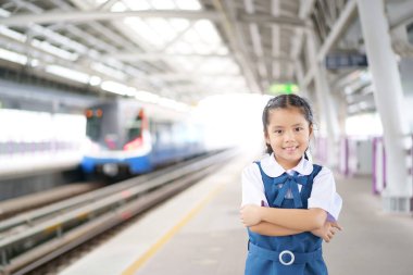 Asyalı çocuk öğrenci mutlu ya da çocuk gülümser. Gökyüzü trenini, tren ve tren yollarını ya da tren istasyonunda metro istasyonunu ya da şehir merkezindeki metro istasyonunu kolları bağlı okul ulaşımına geri dönmek için bekler.