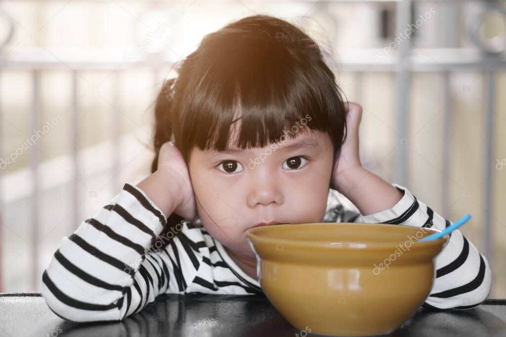 Asian children cute or kid girl student anorexia or sad with vacant and prop up or hand to cheek on food table for breakfast before going to school for study with warm sunlight