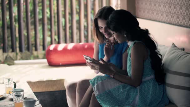 Подружки разговаривают и используют смартфон, сидя на диване — стоковое видео