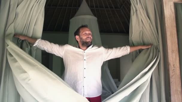 Счастливый человек расстегивает шторы в спальне — стоковое видео