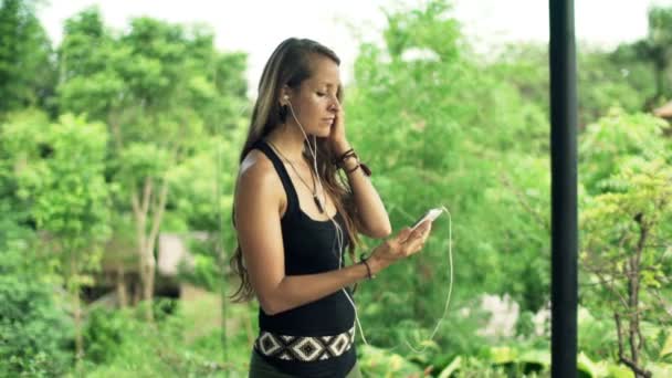 Жінка слухає музику на мобільному телефоні, стоячи в саду — стокове відео