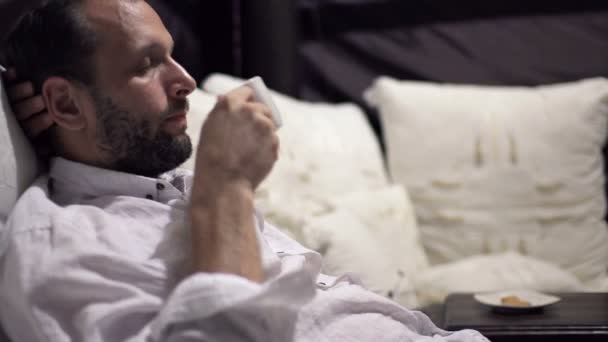 Мужчина пьет кофе и отдыхает на кровати — стоковое видео