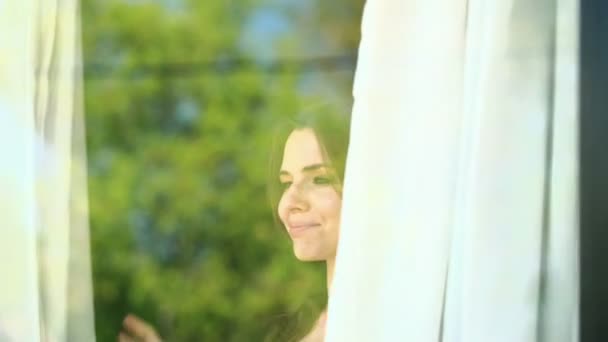 Mujer desvela cortinas, disfrutando de la mañana junto a la ventana — Vídeos de Stock