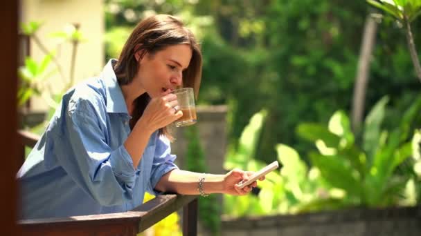 Mujer mensajes de texto en el teléfono inteligente y beber café en la terraza — Vídeos de Stock