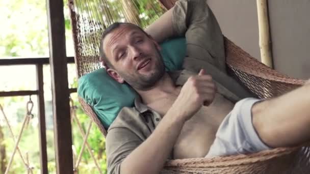 Человек разговаривает с камерой, лежа на гамаке — стоковое видео