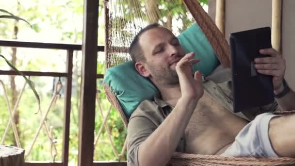 Чоловік балакає на планшетному комп'ютері, лежачи на гамаку — стокове відео