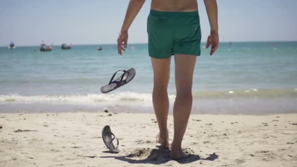 Hombre lanzando flop-flops en la arena y caminando en el mar — Vídeos de Stock