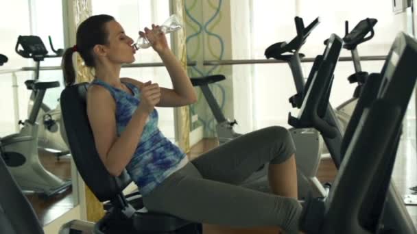 Женщина пьет воду на стационарном велосипеде в тренажерном зале — стоковое видео