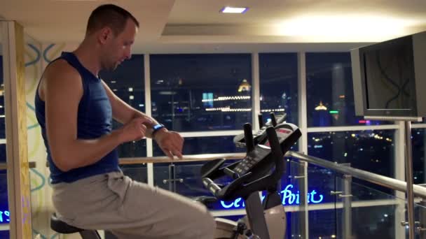 Мужчина с умными часами на стационарном велосипеде в тренажерном зале — стоковое видео