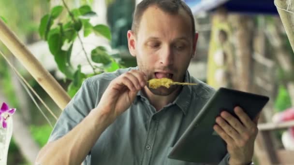 Hombre con el ordenador tableta comer pollo satay — Vídeo de stock