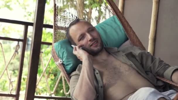 Мужчина разговаривает по телефону, пока лежит на гамаке — стоковое видео