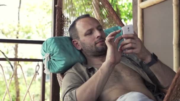 Человек со смартфоном лежит на гамаке — стоковое видео