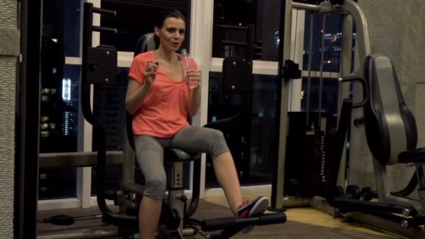 Woman drinking water sitting on machine in gym — Stock Video