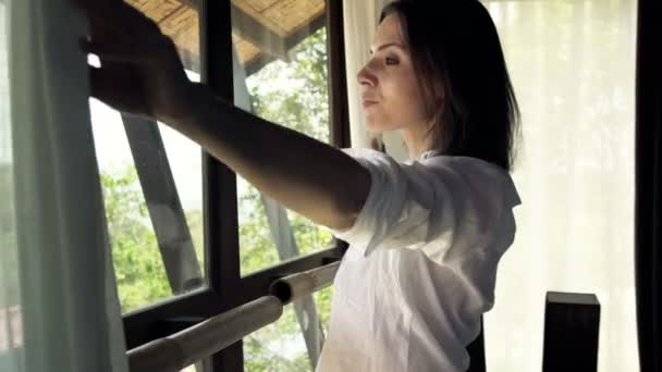 Mujer desvela cortinas, disfrutando de la mañana por la ventana — Vídeos de Stock