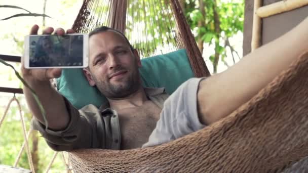 Чоловік фотографує селфі з мобільним телефоном на гамаку — стокове відео
