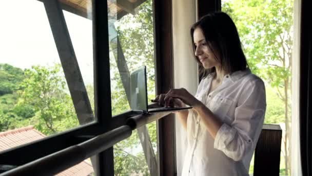Молода жінка, використовуючи ноутбук, що стоїть біля вікна — стокове відео