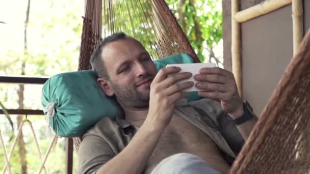 Hombre jugando juego en el teléfono inteligente en la hamaca — Vídeos de Stock