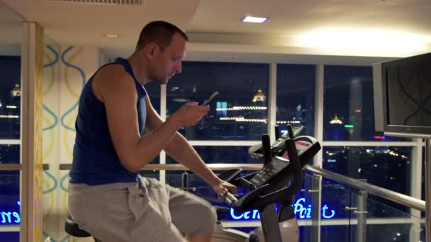 Mann mit Smartphone fährt stationäres Fahrrad in Turnhalle — Stockvideo