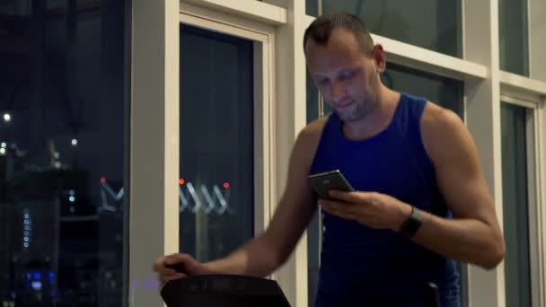 Человек со смартфоном тренируется на эллиптической машине в тренажерном зале — стоковое видео