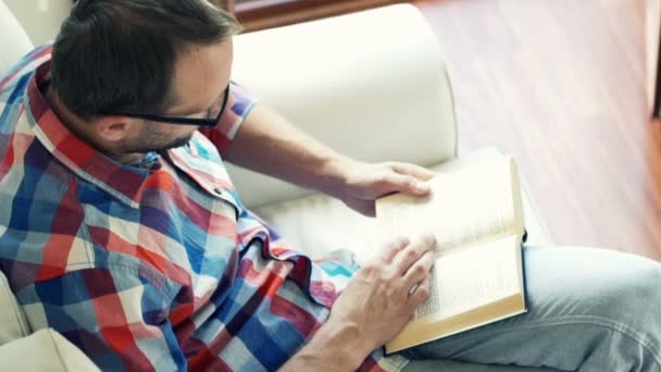 Hombre leyendo libro sentado en sofá en casa — Vídeos de Stock