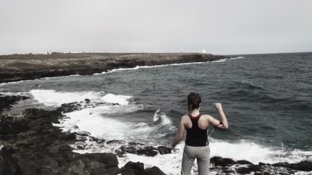 Woman raising arms to the sky standing on rocks by sea — Stock Video