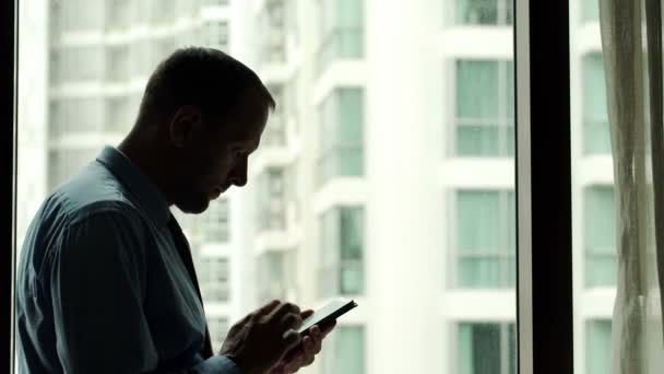 Hombre de negocios con teléfono inteligente de pie junto a la ventana en la oficina — Vídeos de Stock