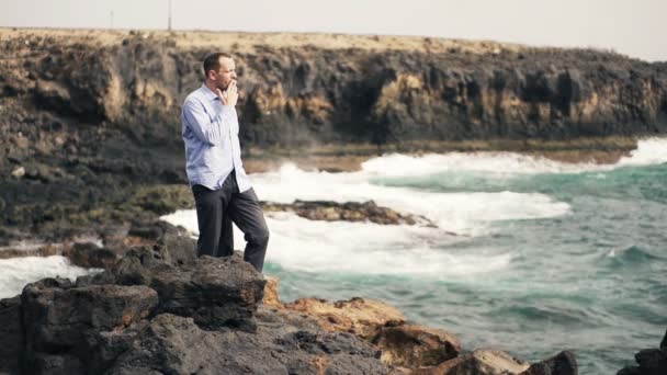 Hombre de negocios fumando cigarrillo en las rocas junto al mar — Vídeos de Stock