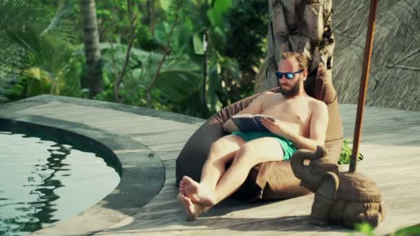 Hombre leyendo libro y relajándose junto a la piscina — Vídeos de Stock