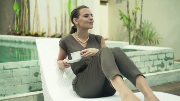 Vrouw drinken koffie zittend op zonnebank bij zwembad — Stockvideo