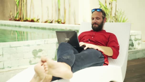 Человек с помощью ноутбука лежит на шезлонге у бассейна — стоковое видео